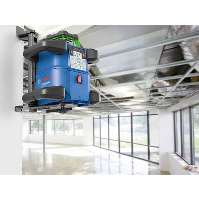 Ein blauer Lasernivellier an einer weißen Wand befestigt, in einem leeren Raum mit Metallträgern an der Decke und großen Fenstern.