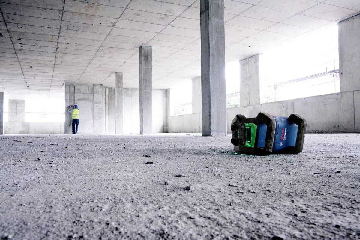 Eine Baustelle mit nacktem Betonboden, Säulen und unvollendetem Dach. Ein Bauarbeiter steht im Hintergrund; ein Werkzeugkasten liegt im Vordergrund.