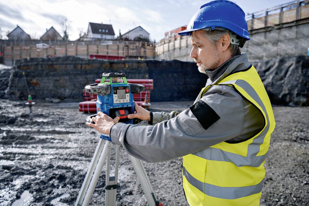 Ein Bauarbeiter in Schutzkleidung und Helm bedient ein Vermessungsgerät auf einer Baustelle. Im Hintergrund sind Erdarbeiten sichtbar.