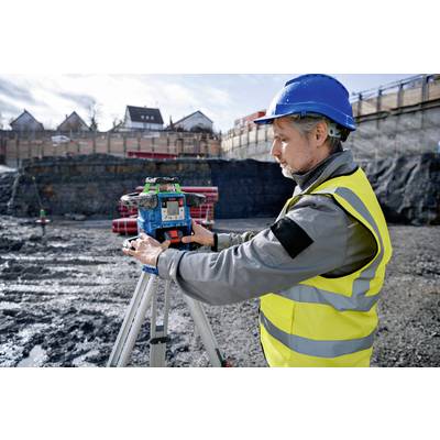 Ein Bauarbeiter in Schutzkleidung und Helm bedient ein Vermessungsgerät auf einer Baustelle. Im Hintergrund sind Erdarbeiten sichtbar.