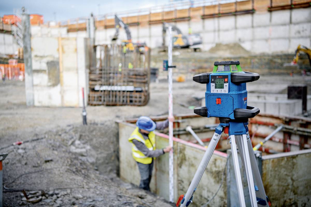 'Eine Baustelle mit einem Laser-Nivelliergerät im Vordergrund. Ein Arbeiter in Schutzkleidung misst in einer Baugrube.'