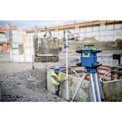 'Eine Baustelle mit einem Laser-Nivelliergerät im Vordergrund. Ein Arbeiter in Schutzkleidung misst in einer Baugrube.'