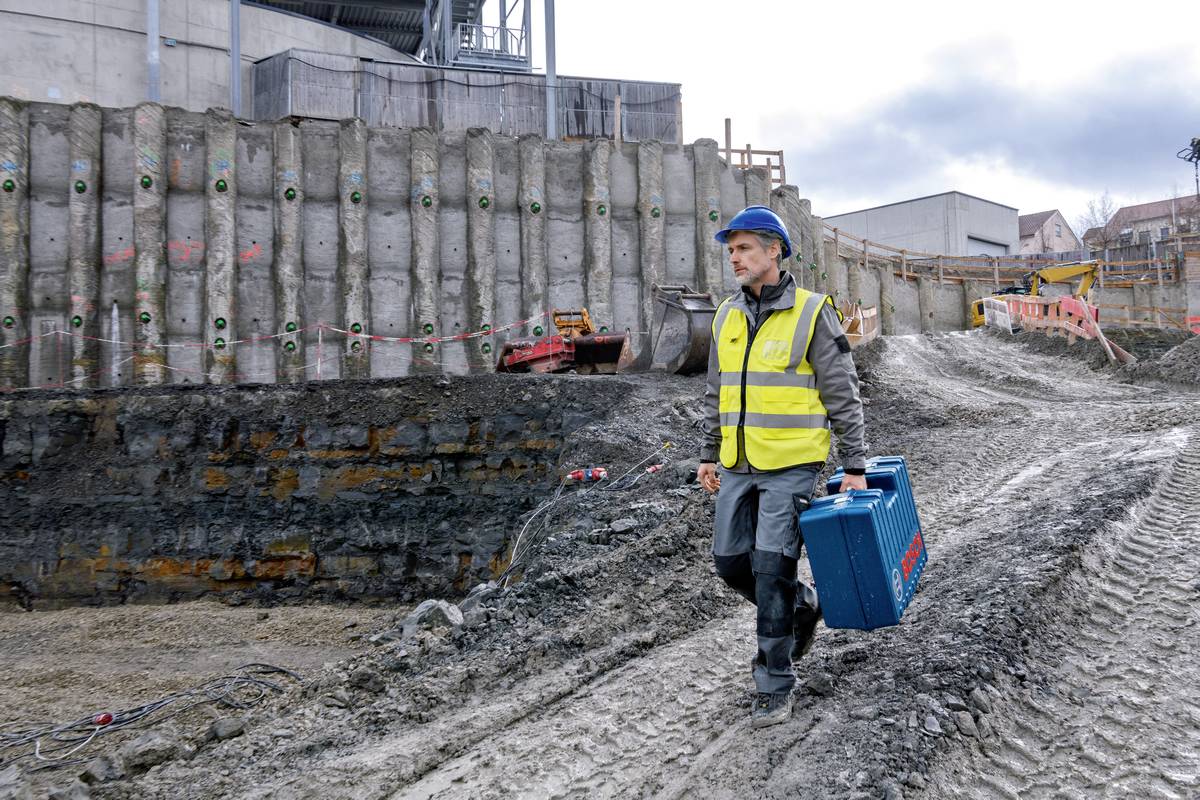 Ein Bauarbeiter in Warnweste und Schutzhelm trägt einen Werkzeugkoffer über eine matschige Baustelle. Im Hintergrund sind Maschinen und Gebäude zu sehen.