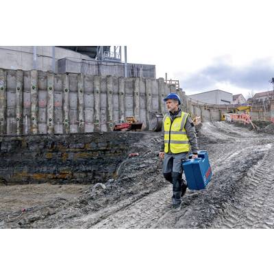 Ein Bauarbeiter in Warnweste und Schutzhelm trägt einen Werkzeugkoffer über eine matschige Baustelle. Im Hintergrund sind Maschinen und Gebäude zu sehen.