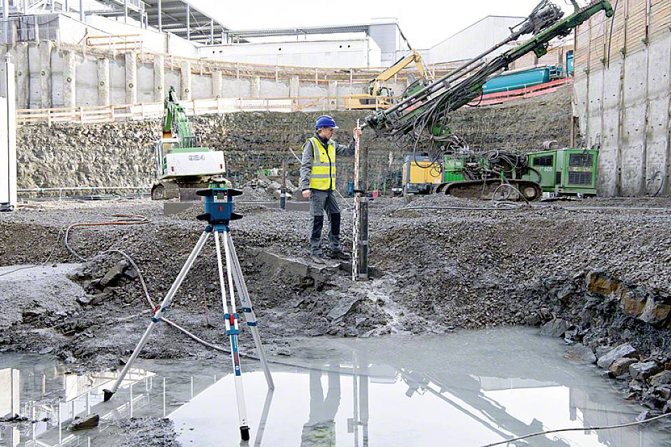 Ein Bauarbeiter in Schutzkleidung bedient schweres Gerät auf einer großen Baustelle mit Erdbohrer und Vermessungsgerät im Vordergrund.
