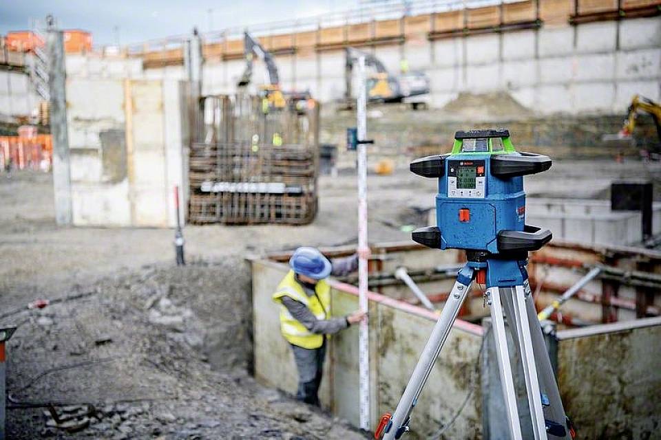 Ein Bauarbeiter mit Helm und Warnweste arbeitet in einer Baugrube, während ein Vermessungsgerät im Vordergrund auf einem Stativ steht.