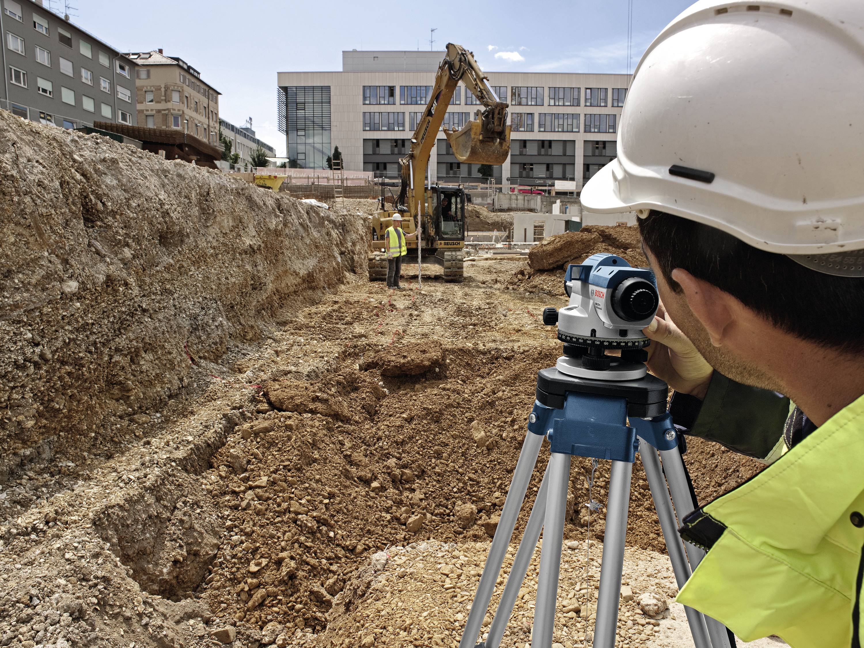Ein Bauarbeiter vermisst eine Baustelle mit einem Vermessungsgerät, während im Hintergrund ein Bagger Erde bewegt.