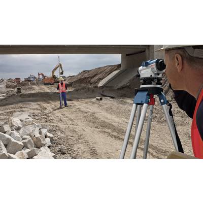 Ein Bauarbeiter verwendet ein Vermessungsgerät, um eine Baustelle unter einer Überführung zu messen. Im Hintergrund sind Bagger zu sehen.