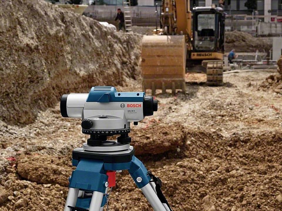 Eine Baustelle mit einem Bau-Nivelliergerät im Vordergrund; im Hintergrund ein Bagger, der Erde bewegt.