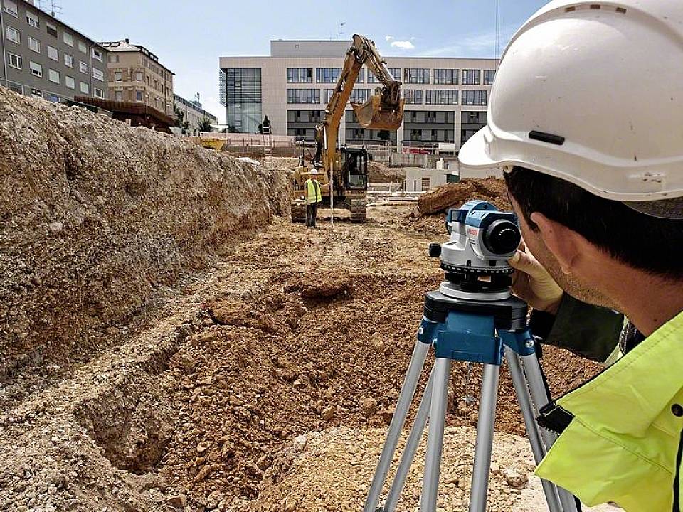 Ein Bauarbeiter mit Helm nutzt ein Vermessungsgerät auf einer Baustelle, um die Erdarbeiten eines arbeitenden Baggers zu überwachen.