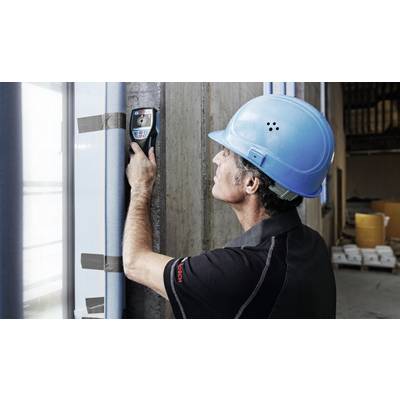 Ein Arbeiter in blauer Schutzhelm und schwarz-grauer Kleidung verwendet ein digitales Messgerät an einer Betonwand in einem industriellen Umfeld.