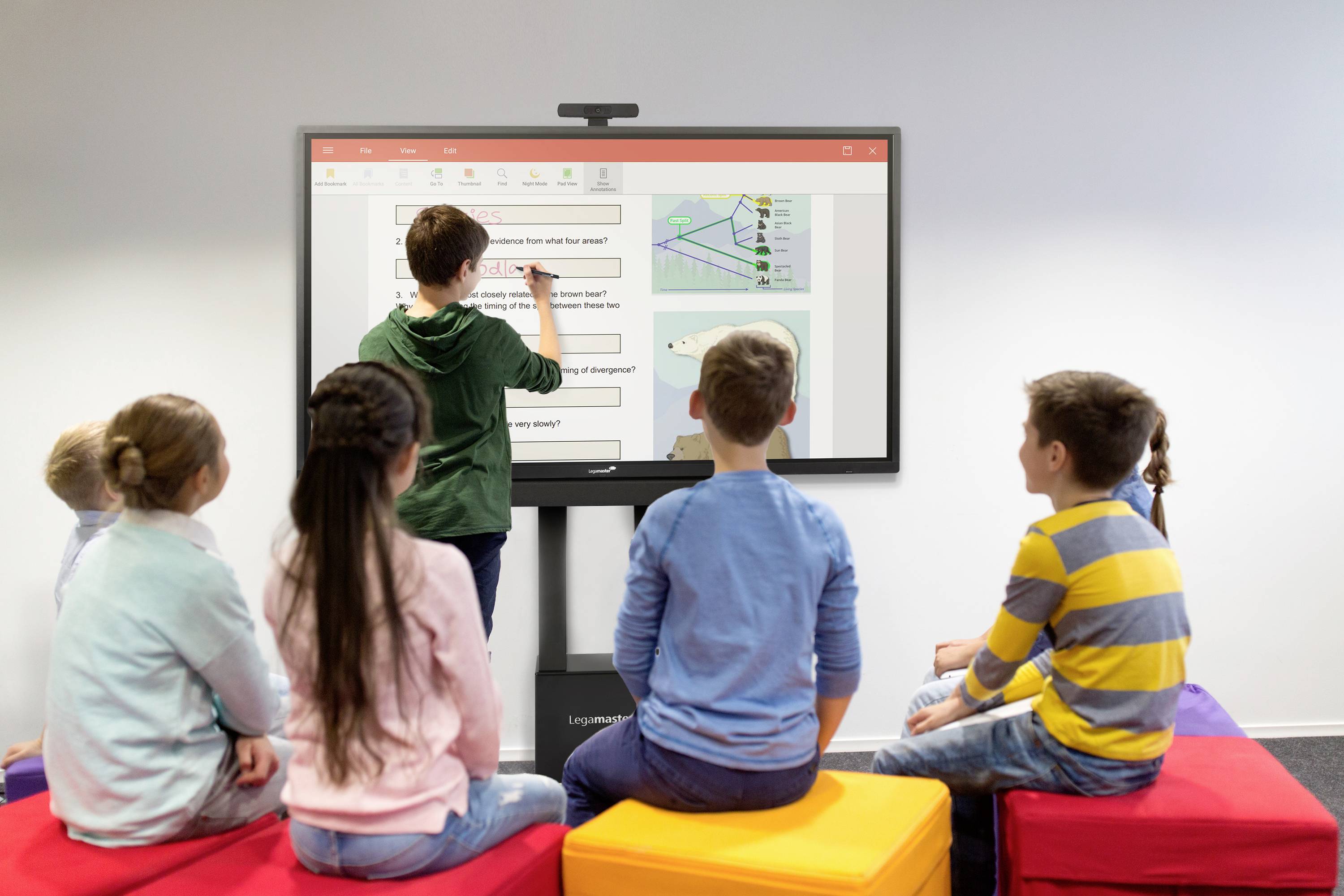 Sechs Kinder sitzen auf bunten Würfeln und schauen auf einen großen Bildschirm. Ein Kind steht und schreibt dort mit einem digitalen Stift.