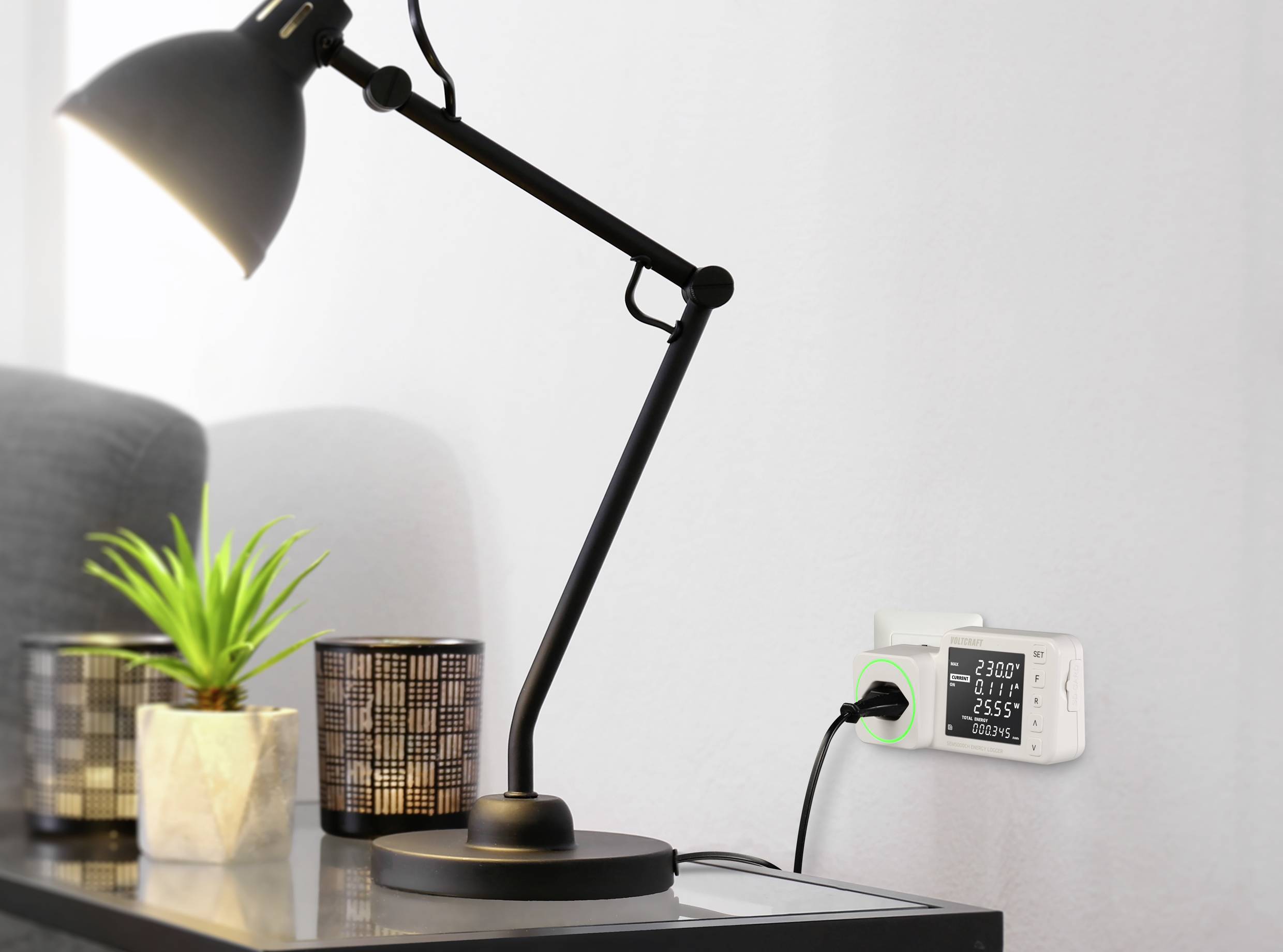Eine Tischlampe steht auf einem Tisch neben einer Pflanze und einem dekorativen Licht. Ein Energiemessgerät zeigt '230 V, 2.55 A'.
