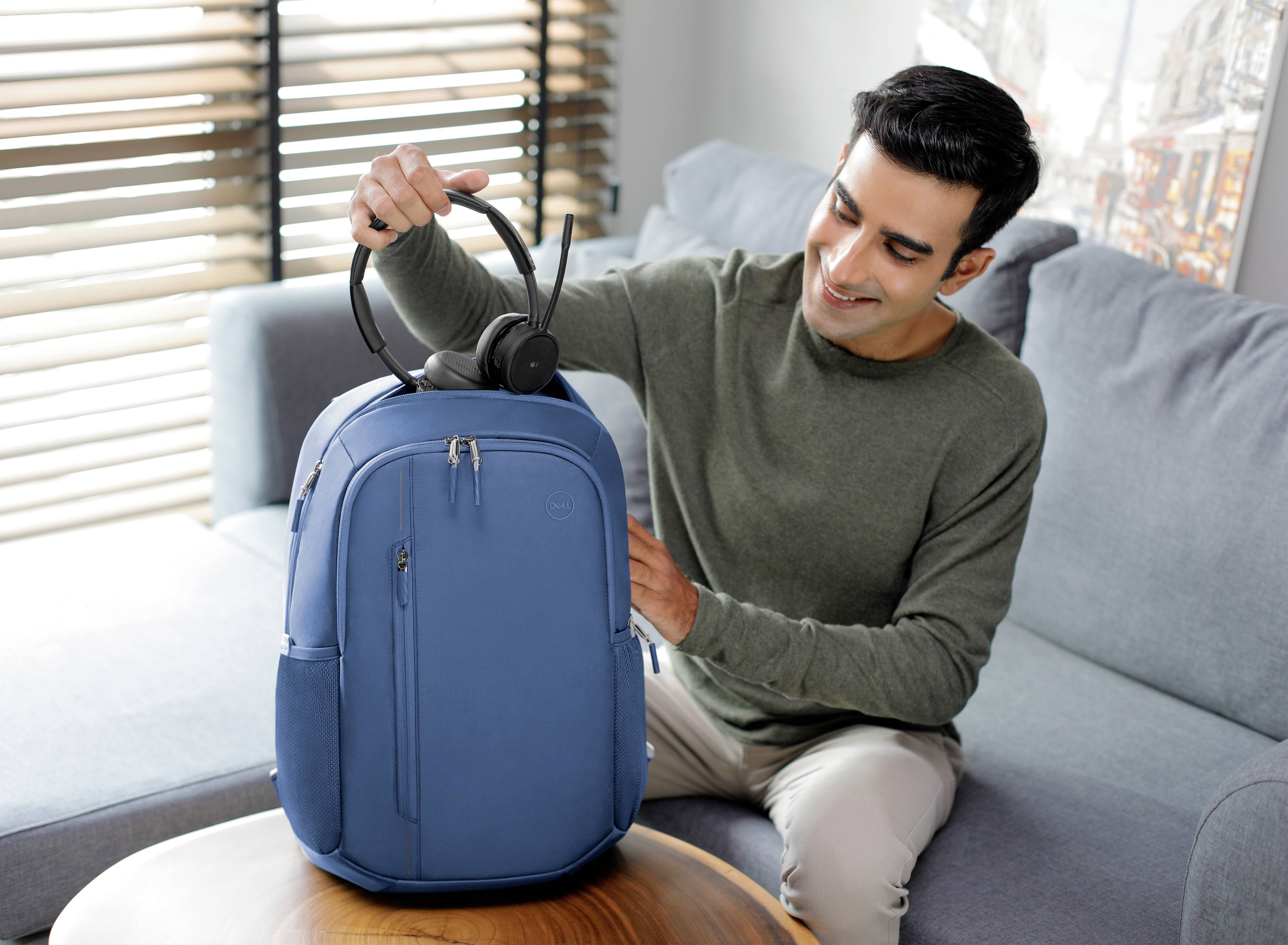 Ein Mann sitzt auf einem Sofa und legt ein Paar Kopfhörer auf einen blauen Rucksack, der auf einem Tisch steht.