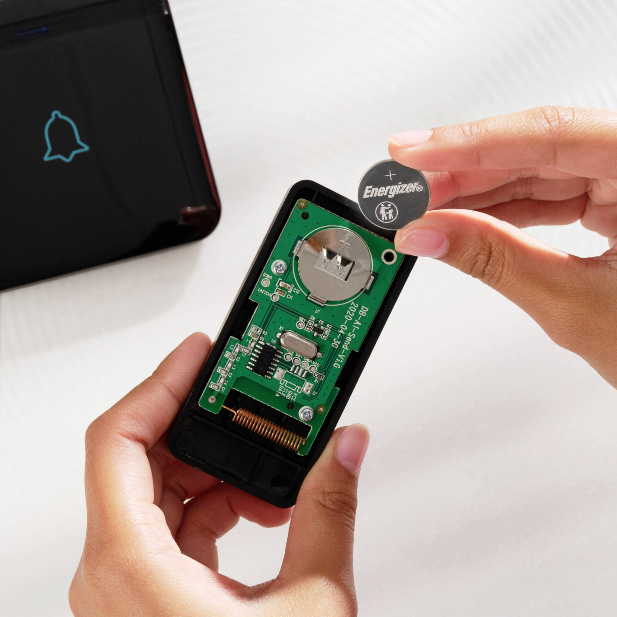 Zwei Hände tauschen eine Batterie in einer elektronischen Fernbedienung aus. Links im Bild ist ein Gerät mit einem Glockensymbol zu sehen.