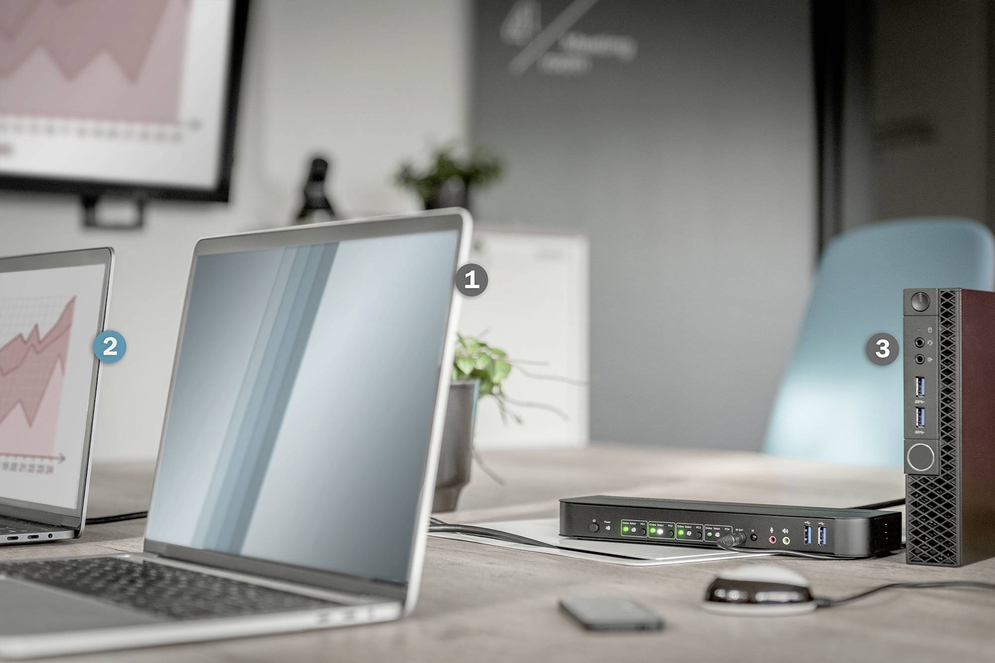 Ein Büro mit zwei geöffneten Laptops auf einem Tisch. Ein Monitor zeigt ein Diagramm. Ein schwarzes Gerät mit Anschlüssen ist sichtbar.