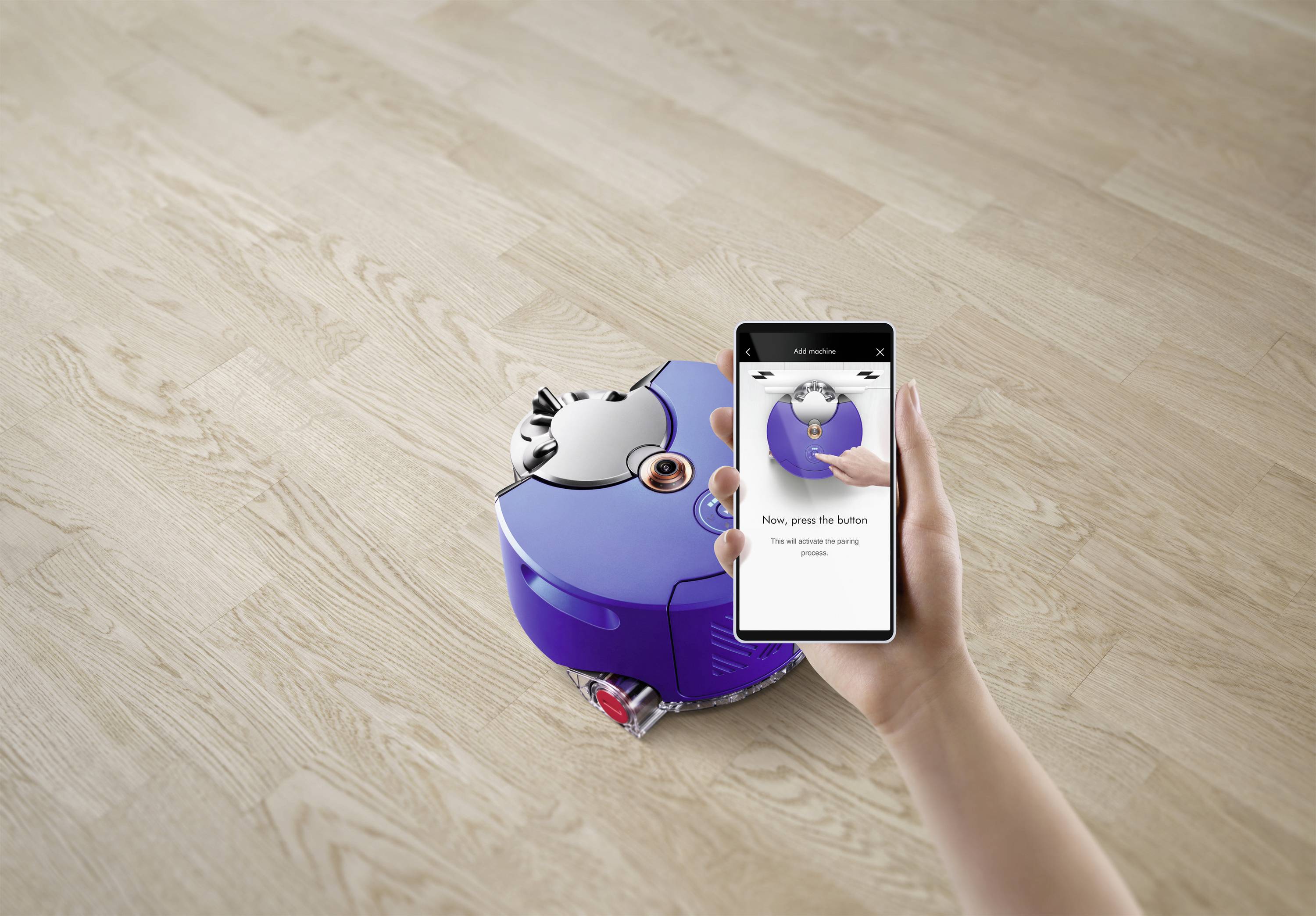 Eine Hand hält ein Smartphone mit einer App, die einen Roboterstaubsauger steuert. Der Roboter saugt auf einem Holzfußboden.