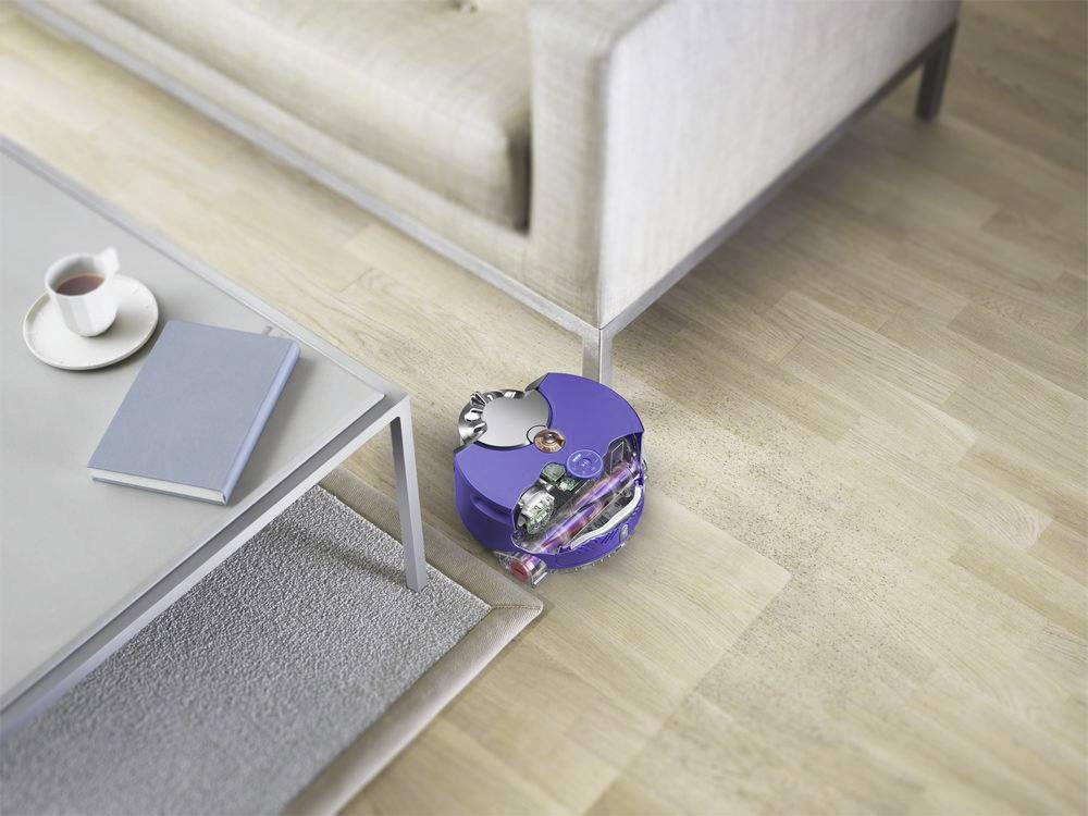Ein runder, violetter Staubsaugerroboter reinigt einen Holzboden neben einem hellgrauen Sofa und einem Tisch mit einem Buch und einer Tasse.