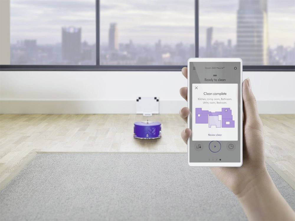 Eine Hand hält ein Smartphone, das einen Plan zur vollständigen Reinigung anzeigt; im Hintergrund ein Roboterstaubsauger in einem Raum.