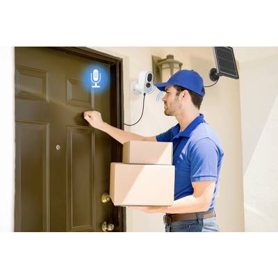 Ein Lieferbote in blauer Uniform klopft an die Tür und hält zwei Pakete. Eine Überwachungskamera mit Mikrofon ist oben montiert.