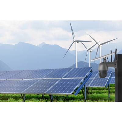 Solaranlagen auf grüner Wiese vor Bergen mit Windkraftanlagen im Hintergrund. Kameraüberwachung im Vordergrund. Thema: erneuerbare Energien.