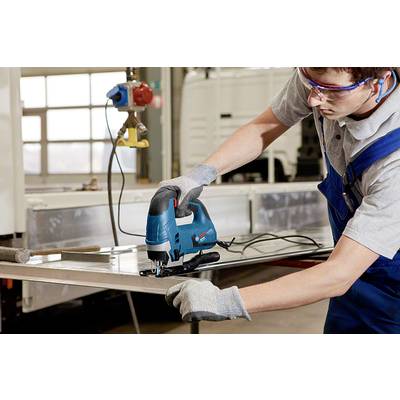 Ein Arbeiter in blauer Latzhose und grauen Handschuhen schneidet mit einer elektrischen Stichsäge ein Stück Metall in einer Werkstatt.