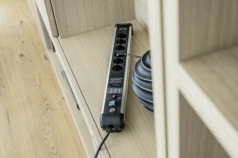 Eine schwarze Steckdosenleiste mit mehreren Anschlüssen liegt auf einem hölzernen Boden. Ein Kabel ist an die Leiste angeschlossen.