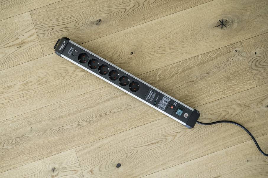 Eine schwarze Mehrfachsteckdose mit sechs Steckplätzen liegt auf einem hellen Holzboden; sie ist eingeschaltet.