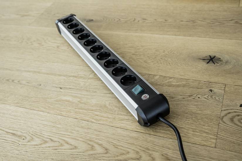 Eine schwarze Mehrfachsteckdosenleiste mit zehn Steckplätzen liegt auf einem Holzboden. Sie ist mit einem Schalter ausgestattet.