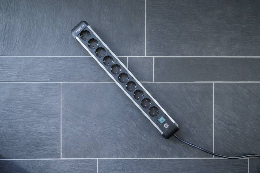 'Steckdosenleiste mit zehn Steckplätzen, auf dunklem Fliesenboden liegend. Ein Kabel führt von der Leiste weg.'