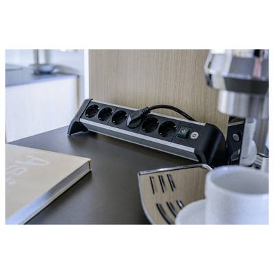 Steckerleiste mit sechs Steckdosen und angeschlossenem Kabel auf einem Schreibtisch neben einer Kaffeemaschine und einer Tasse, was einen Arbeitsbereich oder eine Pausenzone andeutet.