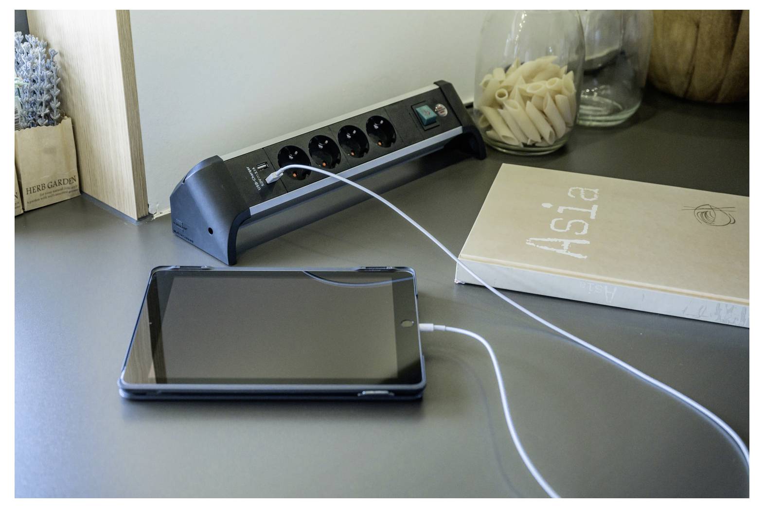 Tablet wird auf einem Schreibtisch neben einer Steckdosenleiste und einem Buch mit dem Titel "Aisla" aufgeladen.