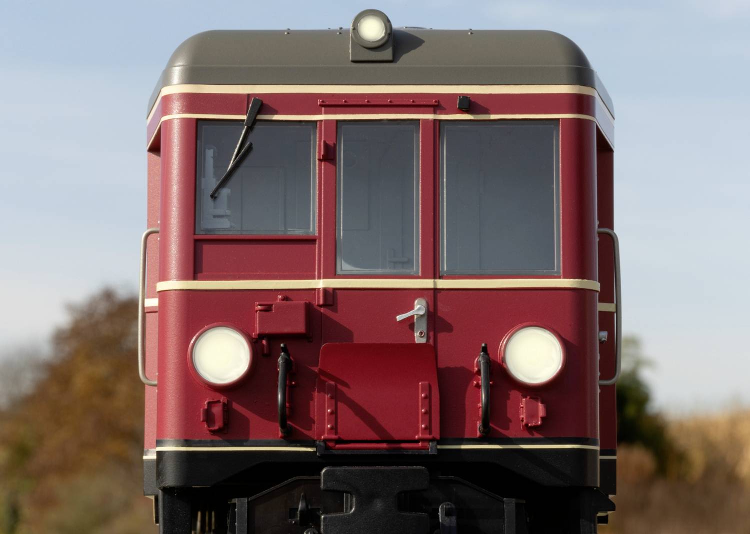 Roter Zug von vorne bei klarem Himmel. Fahrzeug steht still, frontalansicht. Hintergrund zeigt herbstliche Bäume.