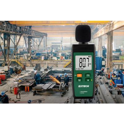 Ein Lärmmessgerät zeigt '80.7 dBA' in einer lauten Fabrikumgebung an. Arbeiter schweißen und bearbeiten Metall.