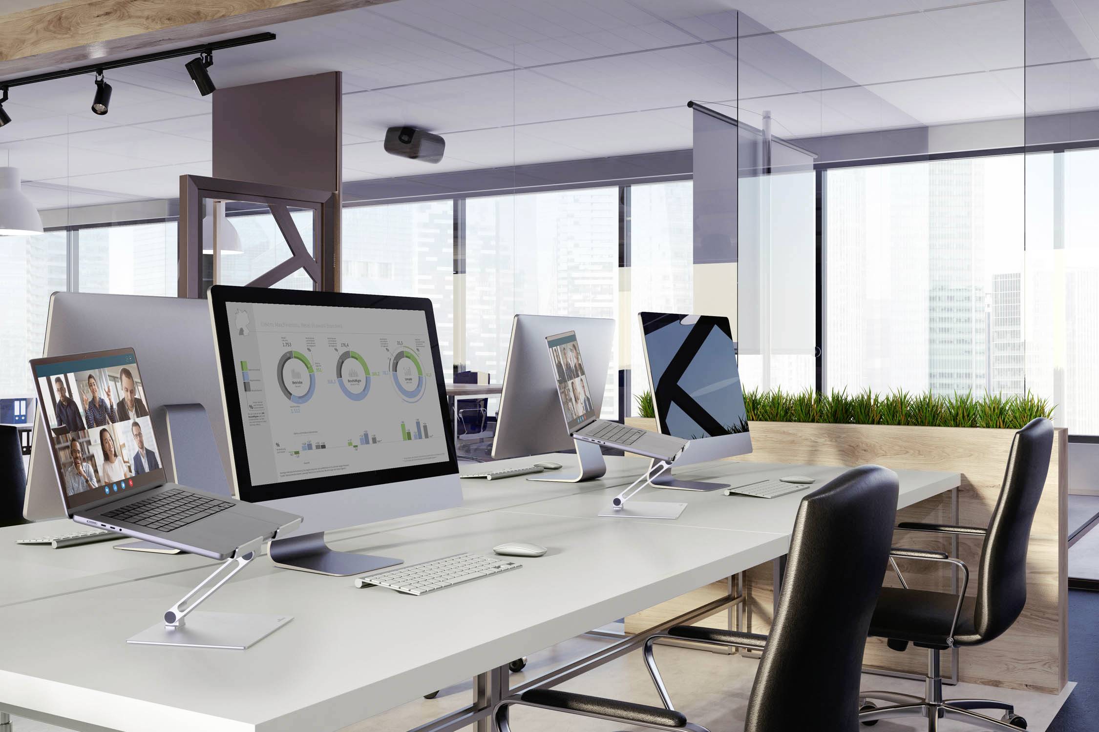 Offenes Büro mit modernen Arbeitsplätzen. Drei Computer zeigen Diagramme und Grafiken. Großes Fenster mit Stadtblick im Hintergrund.