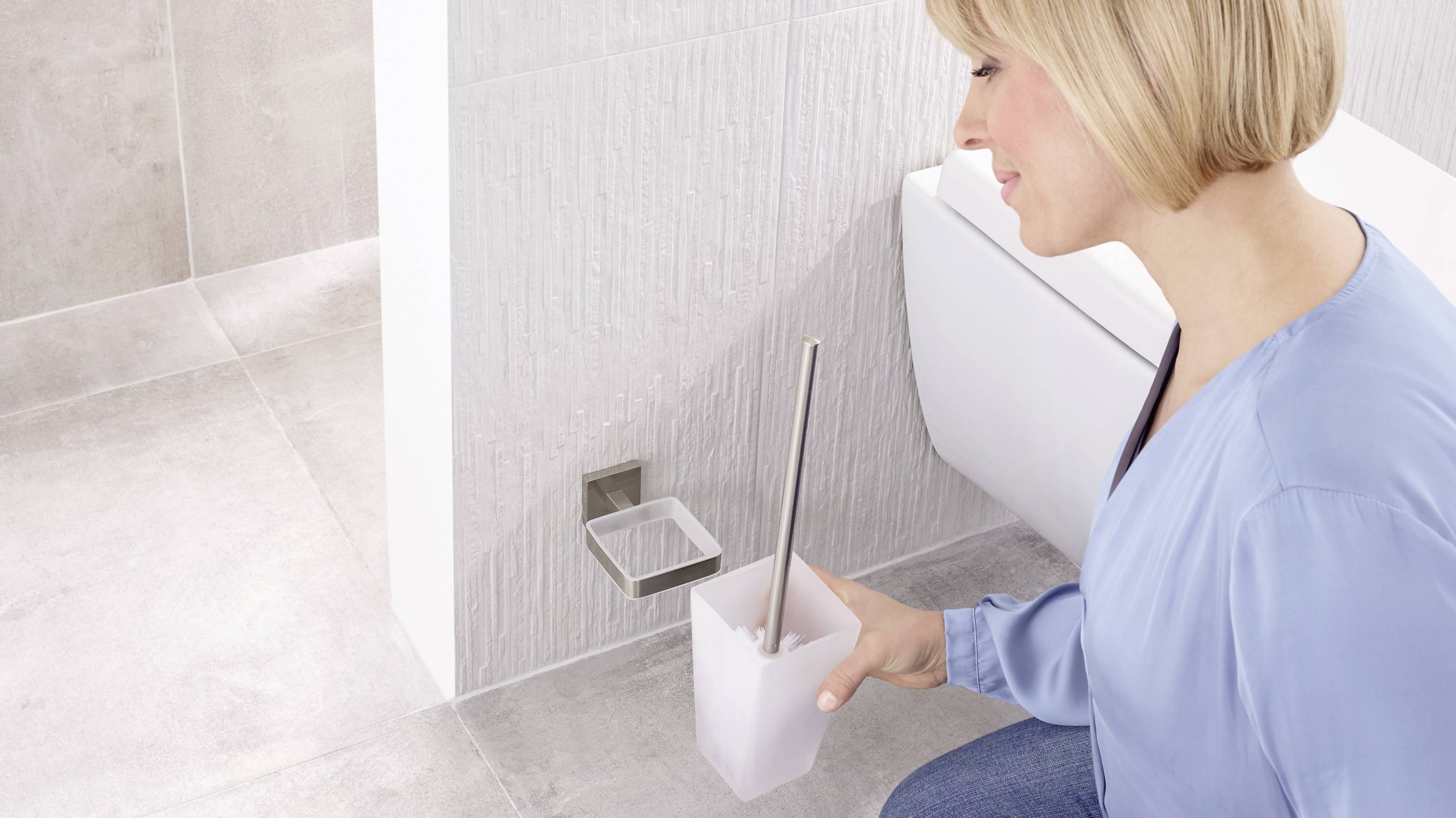 Eine Person reinigt ein Badezimmer, hält eine Toilettenbürste. Im Hintergrund sind Kacheln und ein Teil der Toilette sichtbar.