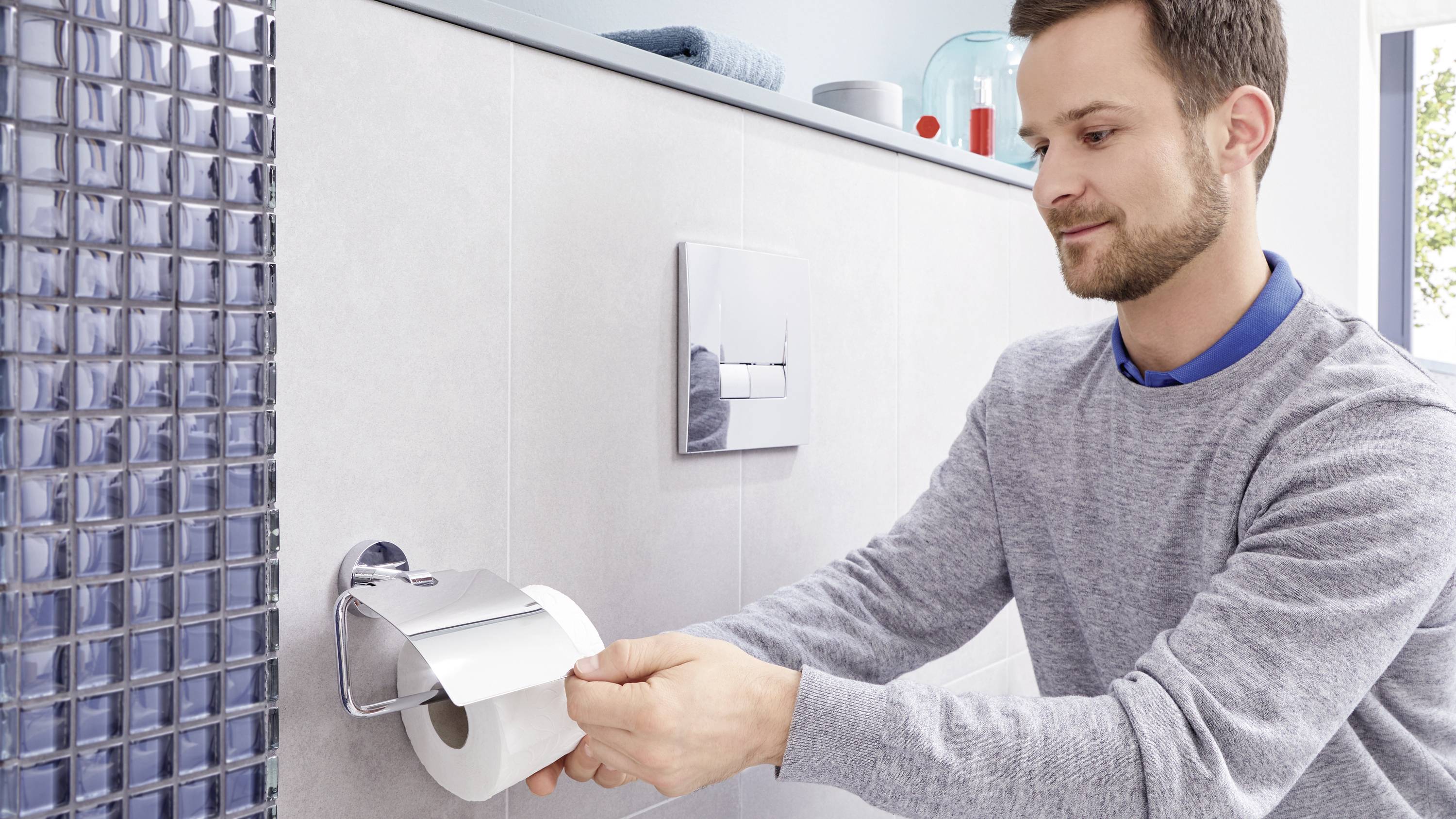 Ein Mann hängt eine Toilettenpapierrolle an einen Halter. Er lächelt. Im Hintergrund sind Badezimmerfliesen und ein Spültastenfeld.