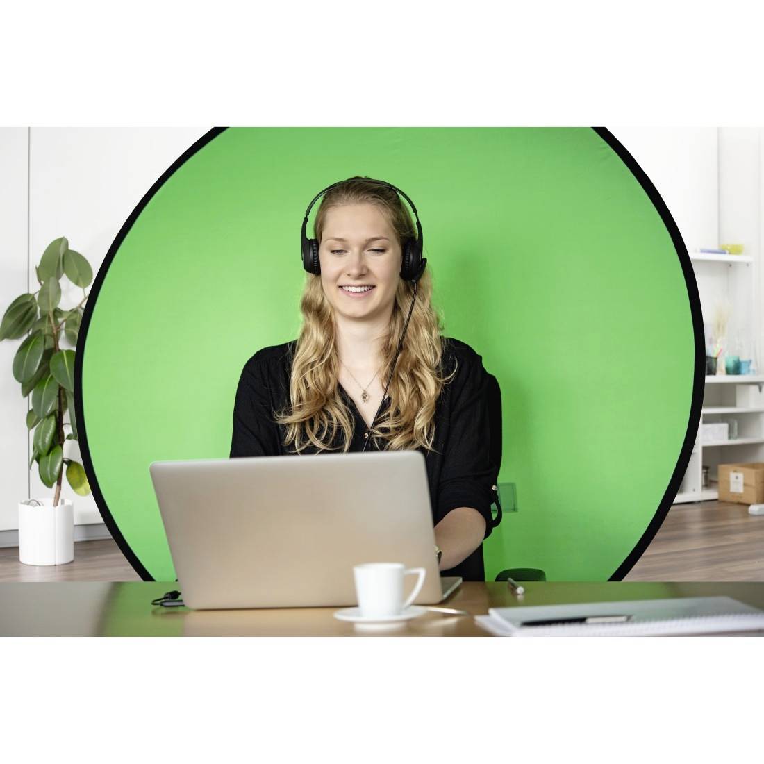 Eine Frau mit Kopfhörern sitzt vor einem Laptop. Im Hintergrund ist ein grüner Bildschirm. Links steht eine Pflanze auf dem Boden.