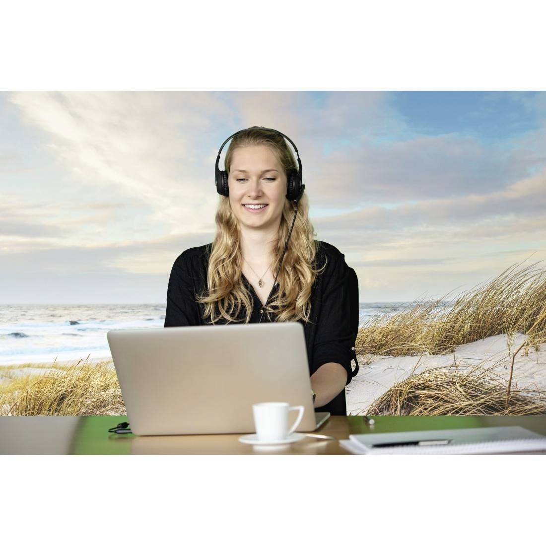 Eine Frau mit Kopfhörern sitzt lächelnd vor einem Laptop. Im Hintergrund ist ein Strand mit Dünen und Meer zu sehen. 