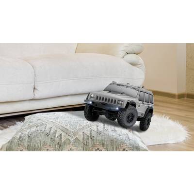 Ein ferngesteuertes Spielzeugauto im SUV-Stil fährt über ein Kissen auf einem hellen Teppich vor einem weißen Sofa in einem Wohnzimmer.