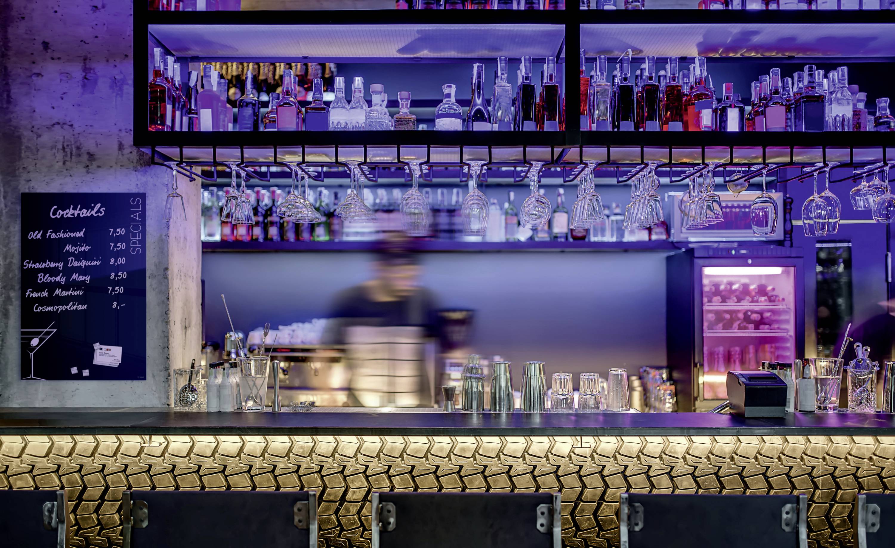 Eine Bar mit beleuchtetem Regal voller Flaschen im Hintergrund. Ein unscharfer Barkeeper bereitet Getränke zu. Eine Tafel zeigt Cocktailpreise.