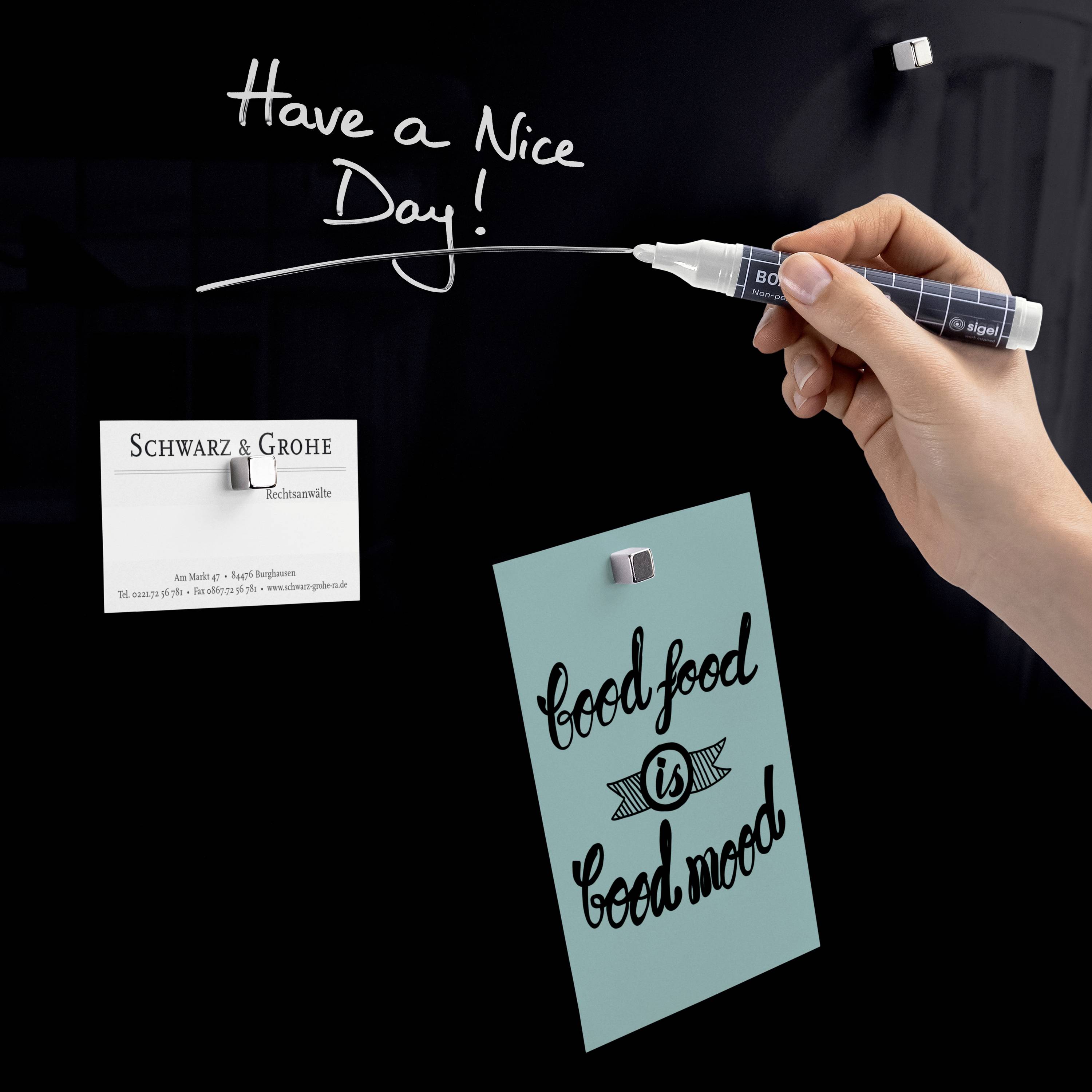 Eine Hand schreibt mit weißem Marker auf Glas 'Have a Nice Day'. Darunter hängt ein blaues Schild mit der Aufschrift 'Good food is good mood'.