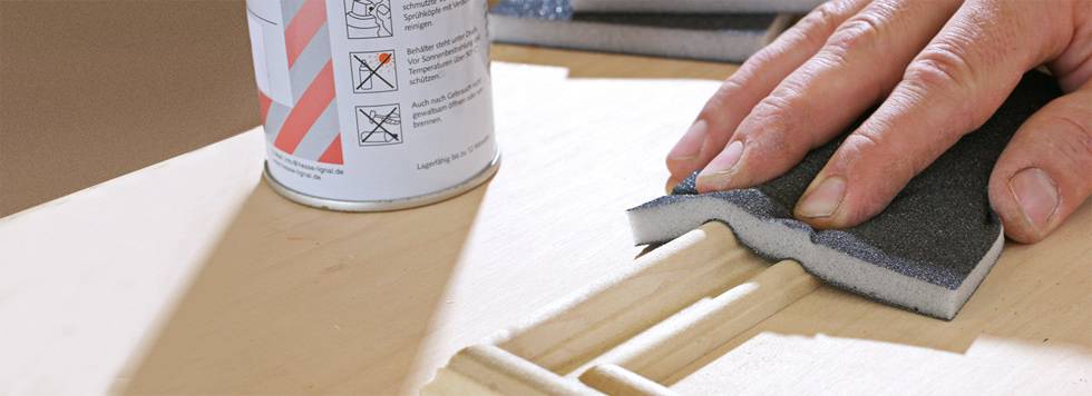 Eine Hand schleift Holz mit einem Schleifschwamm. Neben der Hand steht eine Dose mit Pflegehinweis-Symbolen.