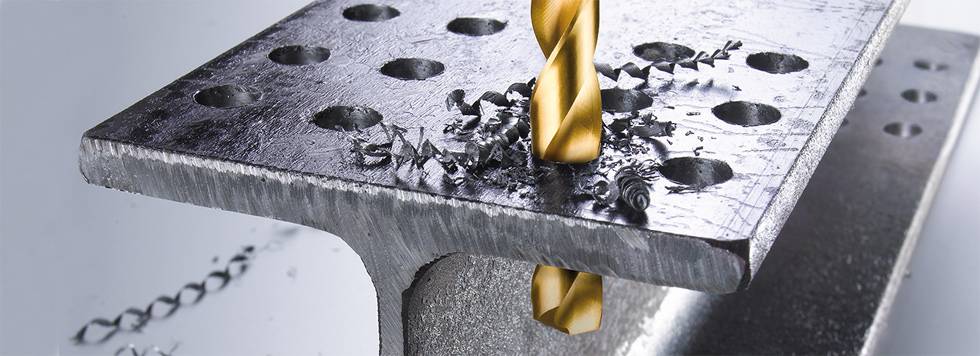 Ein Bohrer durchdringt ein Metallstück mit mehreren vorgebohrten Löchern. Metallspäne sind um die Bohrstelle verteilt.