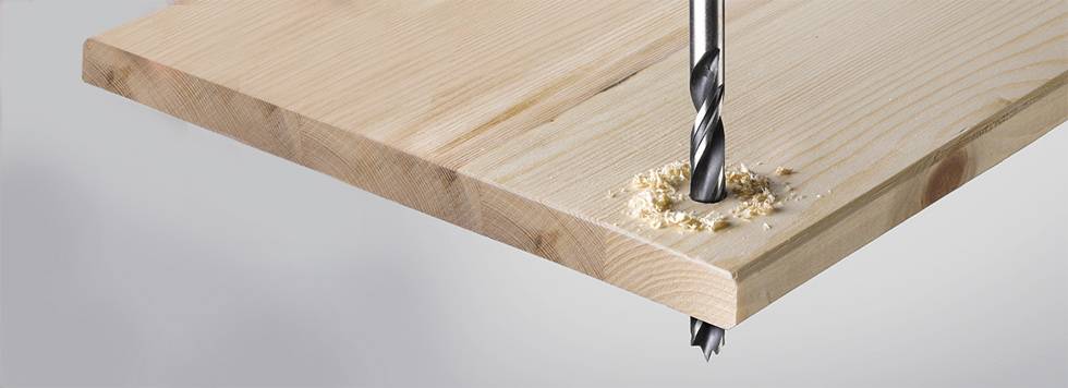 Ein Metallbohrer durchbohrt ein helles Holzbrett, erzeugt Sägespäne.