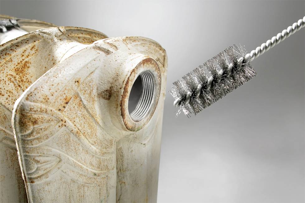 Ein rostiger Metallbehälter mit einer offenen Ausgussöffnung wird mit einer Metalldrahtbürste gereinigt, was auf Wartungs- oder Restaurationsarbeiten hinweist.