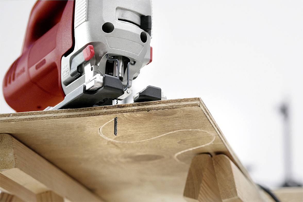 Eine Stichsäge schneidet eine Kurve in eine Holzplatte. Die rote Säge ist im Vordergrund, während die Platte auf Holzböcken liegt.