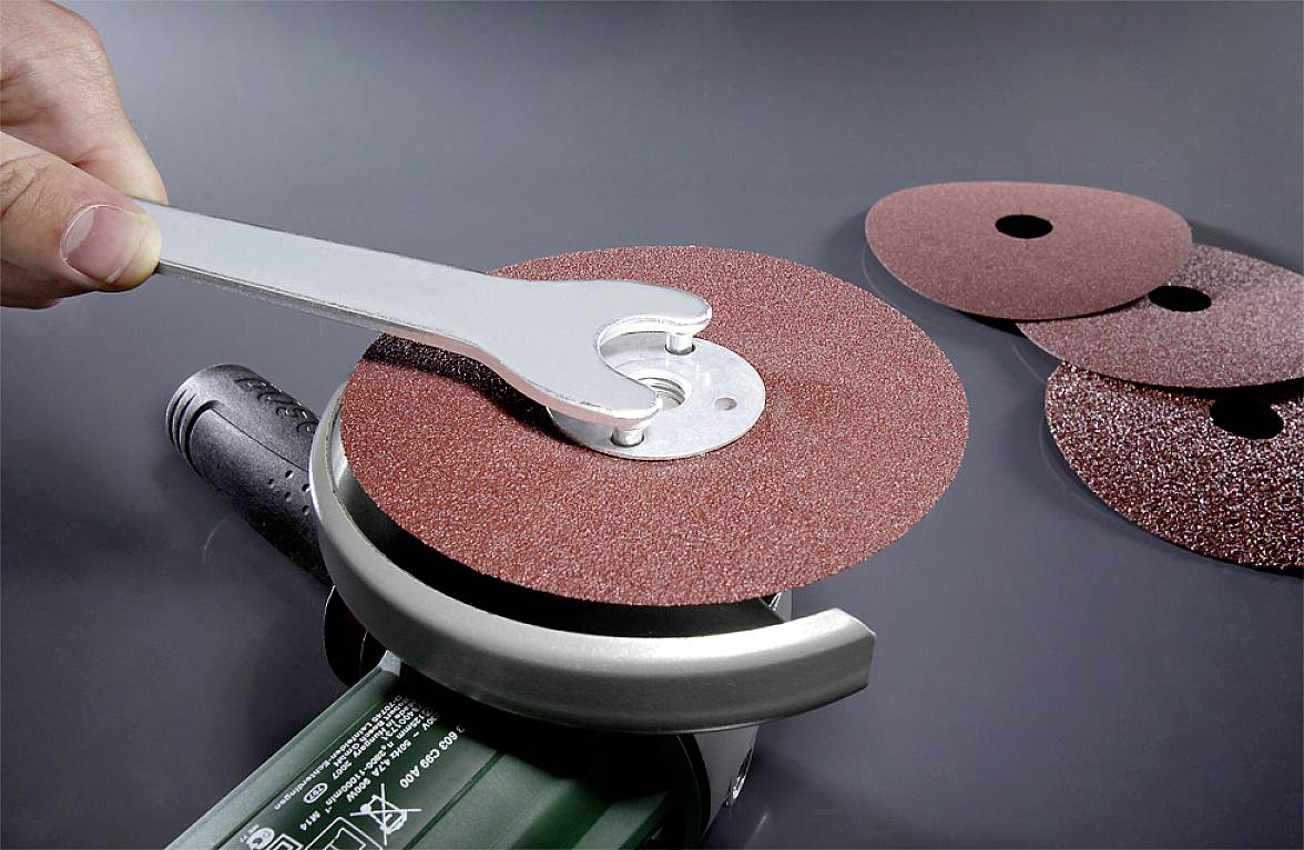 Eine Person befestigt eine Schleifscheibe an einem Winkelschleifer mit einem Schraubenschlüssel. Drei weitere Scheiben liegen daneben.