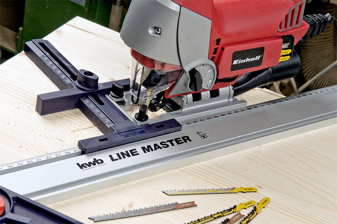 Eine rot-graue Stichsäge auf einem Holzbrett mit Lineal; Sägeblätter und Werkzeuge liegen daneben. Hauptmarke 'Line Master'.