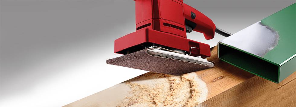 Ein rotes Elektroschleifgerät schleift ein Holzstück. Neben dem Holz liegt ein grünes Metallstück. Fokussiert auf Schleifvorgang.